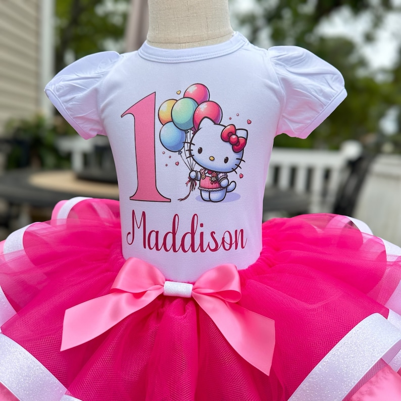 May include: A white t-shirt with a pink number one and a Hello Kitty graphic holding balloons. The shirt has the name Maddison printed below the graphic. The shirt is paired with a pink tulle tutu with a pink satin bow.