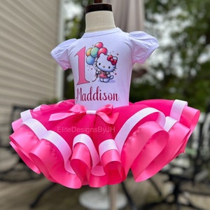 May include: A white t-shirt with a Hello Kitty graphic and the number 1, personalized with the name "Maddison". The shirt is paired with a pink tulle tutu with white satin ribbon accents.
