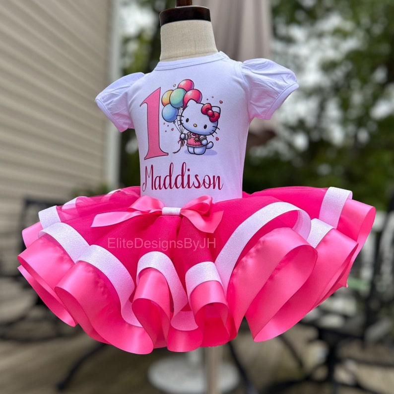 May include: A white t-shirt with a Hello Kitty graphic and the number 1, personalized with the name "Maddison". The shirt is paired with a pink tulle tutu with white satin ribbon accents.