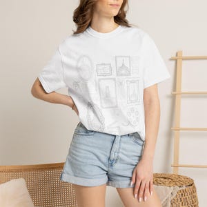 Op de afbeelding: Wit T-shirt met een verzameling ingelijste illustraties, waaronder de Eiffeltoren en een filmrol. Het ontwerp is minimalistisch, met zwarte contouren op een witte achtergrond. Het shirt wordt gedragen met een spijkerbroekshort.