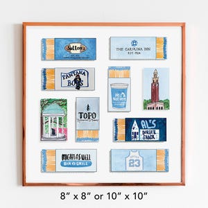 May include: A framed print of nine matchbox labels, each featuring a different design. The labels are all blue and white, and they feature various local businesses and landmarks. The frame is a light brown wood.