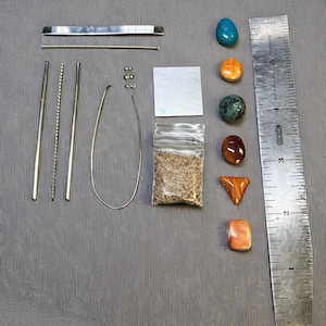 Può includere: Un assortimento di materiali per la creazione di gioielli, tra cui varie aste metalliche, un filo curvo, piccole perline, un quadrato di metallo e un piccolo sacchetto di materiale simile alla sabbia. Sono presenti anche diverse pietre preziose lucide nei toni del blu, arancione, verde e marrone, e un righello di metallo.