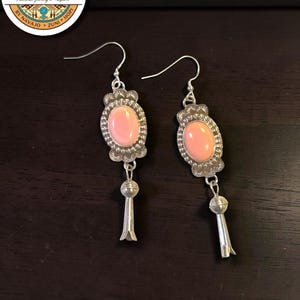 May include: A pair of silver dangle earrings featuring oval-shaped, light pink cabochon stones set in scalloped silver bezels. Each earring includes a silver blossom-shaped drop pendant suspended from the stone setting by a small silver jump ring.