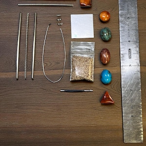 Può includere: Un assortimento di materiali per la creazione di gioielli, tra cui barre di metallo, perline, un sacchetto trasparente con materiale simile alla sabbia e varie pietre preziose lucide nei toni del marrone, arancione, blu e verde. È presente anche un righello di metallo.