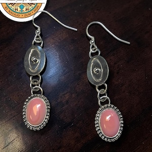 May include: A pair of silver drop earrings featuring a polished oval pink stone set in a beaded frame, suspended from a smaller oval silver disc with a central stud detail. The earrings include silver hook wires for pierced ears.