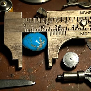 Può includere: Una pietra preziosa turchese viene misurata con un calibro metallico. La pietra è di un blu vibrante con inclusioni color argento. Il calibro mostra una misura di circa 1,9 cm. Vari strumenti e componenti circondano la pietra.