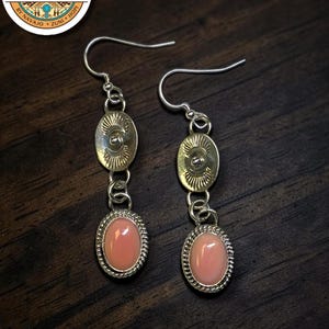May include: A pair of silver drop earrings featuring a polished oval pink stone set in a twisted silver border, suspended from a smaller oval silver disc with sunburst engraving and a central stud, attached to silver ear wires.