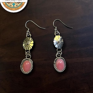 May include: A pair of silver drop earrings featuring a circular sunburst-style metal charm connected to an oval-shaped pink cabochon stone set in a beaded silver frame. The earrings include silver fishhook wires for attachment to the ear.