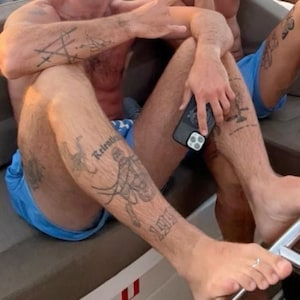 Peut inclure: Une personne portant un short bleu avec des tatouages sur les bras et les jambes. Les tatouages comprennent des formes géométriques, du texte et des illustrations. Il tient un smartphone noir avec un appareil photo à trois objectifs.