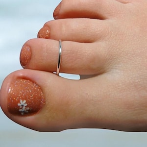 May include: A silver toe ring on a foot with a glittery orange toenail with white flower designs.