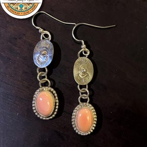 May include: A pair of silver drop earrings featuring an oval-shaped, light pink cabochon stone set in a beaded bezel. Above the stone, a circular silver disc displays a stamped sunburst pattern. The earrings hang from simple silver ear wires.