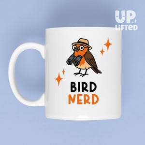 May include: White ceramic mug with a cartoon bird wearing a hat and binoculars. The bird is orange, brown, and black. The mug has the words "BIRD NERD" in black and orange. The background is light blue.