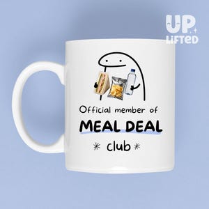 May include: A white ceramic mug featuring a black line-art cartoon character holding a sandwich, a bag of chips, and a water bottle. Below the character, the text reads: Official member of MEAL DEAL club, with the words MEAL DEAL in bold blue.
