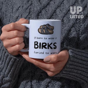 Peut inclure: Mug en c&eacute;ramique blanche avec le texte "born to wear BIRKS forced to work" et un dessin de sandales. La tasse est tenue par une personne portant un pull gris. La marque "Up Lifted" est dans le coin sup&eacute;rieur droit.
