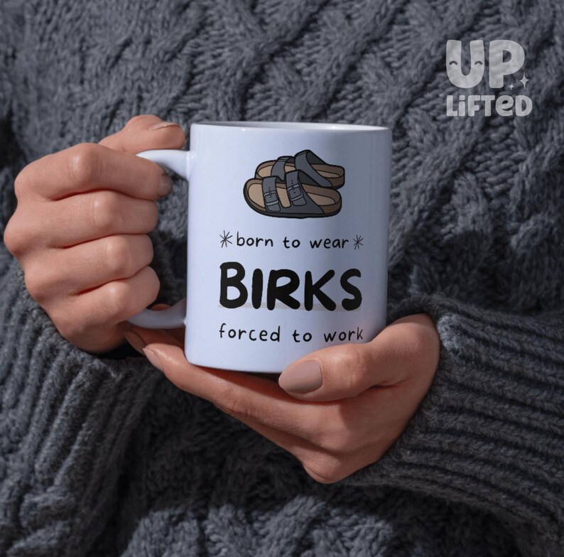 Peut inclure: Mug en c&eacute;ramique blanche avec le texte "born to wear BIRKS forced to work" et un dessin de sandales. La tasse est tenue par une personne portant un pull gris. La marque "Up Lifted" est dans le coin sup&eacute;rieur droit.