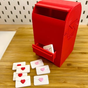 May include: A red mailbox with a drawer holding a white envelope with a pink heart. Several white envelopes with red and pink hearts are scattered around. The mailbox features an embossed envelope design.