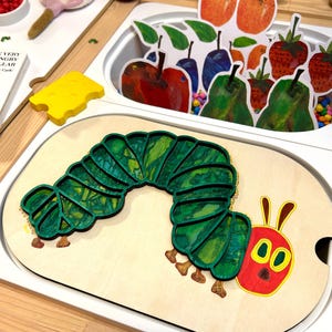 May include: A wooden puzzle depicting a green caterpillar with a red head and yellow eyes. The puzzle sits on a light wooden base, surrounded by colourful fruit cutouts. The Very Hungry Caterpillar theme is visible.