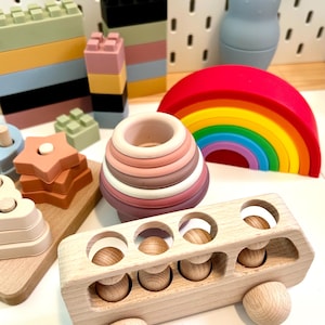 May include: A collection of colorful wooden and silicone toys, including a rainbow, stacking blocks, a hippo, and a bus with wooden pegs. The toys are in various pastel colors, designed for children's play and development.