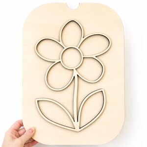 May include: A wooden flower cutout with a light brown finish. The flower has five petals, a circular centre, a stem, and two leaves. The cutout is held by a hand against a white background.