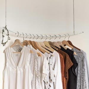 May include: A clothing rack made from a white branch, suspended from the ceiling, holds a variety of garments on wooden hangers. The clothes include white, patterned, and solid-colored tops and a gray cardigan. A decorative garland hangs from the branch.