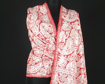 Puede incluir: Un chal rojo con bordados blancos en forma de paisley. El chal tiene un borde de flecos y está drapeado sobre un maniquí.