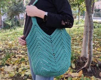 May include: A teal crocheted tote bag with a chevron pattern. The bag is being worn by a person with a black long-sleeved shirt and blue jeans.
