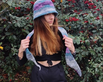 May include: A woman wearing a blue, purple, and gray striped crocheted hat and matching scarf. The hat is a cloche style with a wide brim. The scarf is a triangular shape and is draped around her neck.