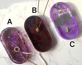 Könnte beinhalten: Drei ovale Schalen aus Harz mit einer violetten und glitzernden Oberfläche. Die Schalen sind mit Glitzer gefüllt und haben verschiedene Designs. Eine Schale hat einen goldenen Augen-Anhänger, eine andere einen goldenen Sonnen-Anhänger und die dritte einen Schmetterlings-Anhänger.