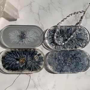 Può includere: Quattro vassoi ovali in resina con un design floreale. I vassoi sono trasparenti e hanno un design floreale bianco e nero con accenti di glitter. Un vassoio ha una collana di perle sopra.