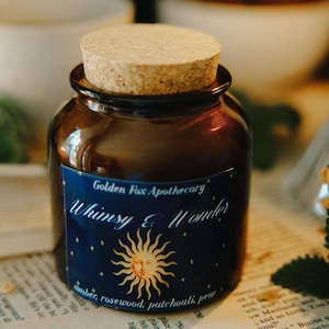 May include: A small, brown apothecary jar with a cork stopper, labeled "Whimsy & Wonder" with a sun and star design. The jar sits on a vintage book page, with a teacup and candle in the background. The label lists amber, rosewood, and patchouli.