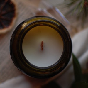 Op de afbeelding: Bovenaanzicht van een kaars in een bruine glazen pot. De kaars heeft witte was en een houten lont. Gedroogde sinaasappelschijfjes en groen in de achtergrond cre&euml;ren een natuurlijke, rustieke uitstraling.