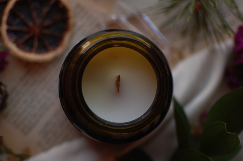 Op de afbeelding: Bovenaanzicht van een kaars in een bruine glazen pot. De kaars heeft witte was en een houten lont. Gedroogde sinaasappelschijfjes en groen in de achtergrond cre&euml;ren een natuurlijke, rustieke uitstraling.