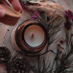 Op de afbeelding: Een bruine glazen kaarsenpot met een witte kaars en een houten lont. De pot wordt afgedekt met een kurken deksel. De sc&egrave;ne bevat dennennaalden, dennenappels en gedroogde bloemen, wat een natuurlijke, rustieke esthetiek cre&euml;ert.