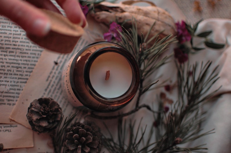 Op de afbeelding: Een bruine glazen kaarsenpot met een witte kaars en een houten lont. De pot wordt afgedekt met een kurken deksel. De sc&egrave;ne bevat dennennaalden, dennenappels en gedroogde bloemen, wat een natuurlijke, rustieke esthetiek cre&euml;ert.