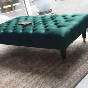 May include: A dark green velvet ottoman with tufted buttons and gold-coloured legs. The ottoman is sitting on a rug with a light brown and beige pattern.