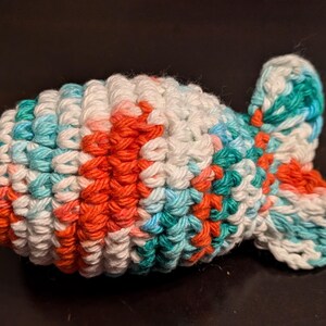 May include: A crocheted fish toy with a white, teal, and orange color scheme. The fish has a rounded body and a small tail fin.