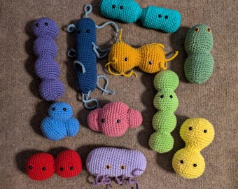 Puede incluir: Una colección de doce coloridos peluches de ganchillo con formas de diversos microorganismos. El conjunto incluye figuras de color morado, azul, turquesa, amarillo, verde, rosa, rojo y morado claro, cada una con pequeños ojos de cuentas negras.