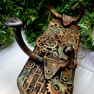 May include: A steampunk-style wall hook with a dark, aged finish. The hook features intricate gear and cog details, with a combination of bronze, copper, and teal colors. The hook is designed to hold items and adds a unique, industrial touch to any space.