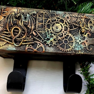 May include: A steampunk-themed wall shelf with a dragon and gear design. The shelf is made of wood and features a detailed relief of a dragon, gears, and other mechanical elements. The color palette includes bronze, copper, and teal accents. Two black hooks are attached to the bottom.