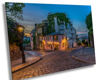 May include: A cobblestone street in Paris, France, with a pink building and a street lamp. The street is lit by the lamp and the building is covered in ivy.