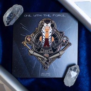 May include: A gold and black enamel pin featuring Ahsoka Tano in a meditative pose. The pin is set against a dark blue background with a starry sky and the text "One with the Force".