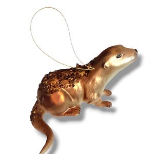 May include: A brown otter ornament with a smooth finish on the head and limbs, featuring a textured back covered in bronze-colored glitter. The figure includes a thin gold hanging cord attached to the top, designed for display as a hanging decoration.