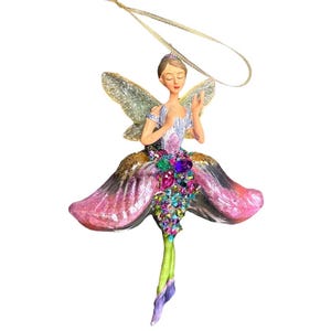 May include: A decorative fairy ornament with a serene expression. The fairy has glittery wings and a dress made of pink and purple petals, adorned with colorful jewels. A gold ribbon is attached for hanging.