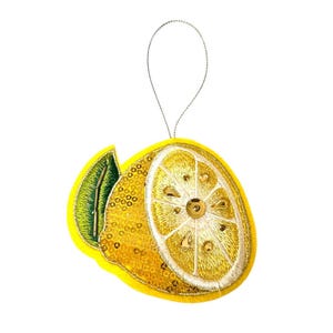 Könnte beinhalten: Ein gelber, zitronenförmiger Schmuckanhänger mit einer silbernen Schnur zum Aufhängen. Der Anhänger ist mit goldenem Faden und Pailletten bestickt und hat ein grünes Blatt. Die Zitrone ist halbiert und zeigt die inneren Details.