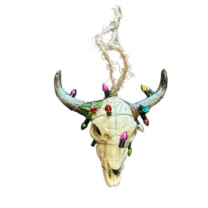 May include: A white bull skull Christmas ornament with a string of colorful lights wrapped around it. The skull has a rustic, weathered look and is decorated with green holly leaves and red berries.