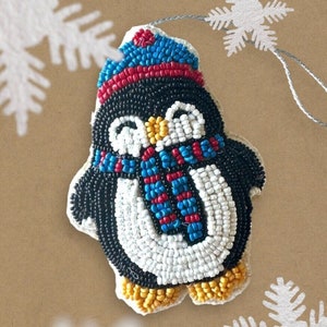 Pode incluir: Um enfeite de Natal de pinguim com contas. O pinguim é preto, branco e amarelo, com um chapéu e cachecol azuis e vermelhos. O enfeite é enfiado com um cordão prateado e tem um floco de neve.