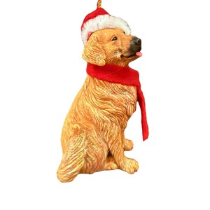 May include: A golden retriever figurine wearing a red and white Santa hat and a red scarf. The dog is sitting and has its tongue sticking out. The figurine is a Christmas ornament.