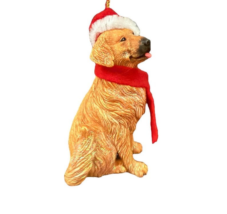 May include: A golden retriever figurine wearing a red and white Santa hat and a red scarf. The dog is sitting and has its tongue sticking out. The figurine is a Christmas ornament.