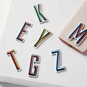 Op de afbeelding: Kleurrijke alfabetsticker in verschillende kleuren, waaronder rood, blauw, groen, geel en wit. De letters zijn gerangschikt op een wit oppervlak en omvatten de letters K, E, Y, T, G, Z en M.