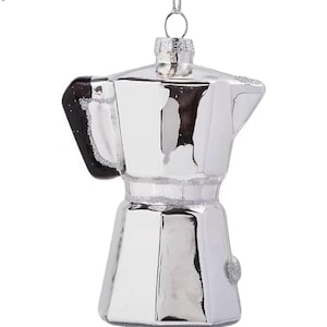 Puede incluir: Un adorno navideño plateado con forma de cafetera italiana. El adorno tiene un asa negra y un lazo plateado para colgar. El adorno es de vidrio y tiene una superficie brillante y reflectante.